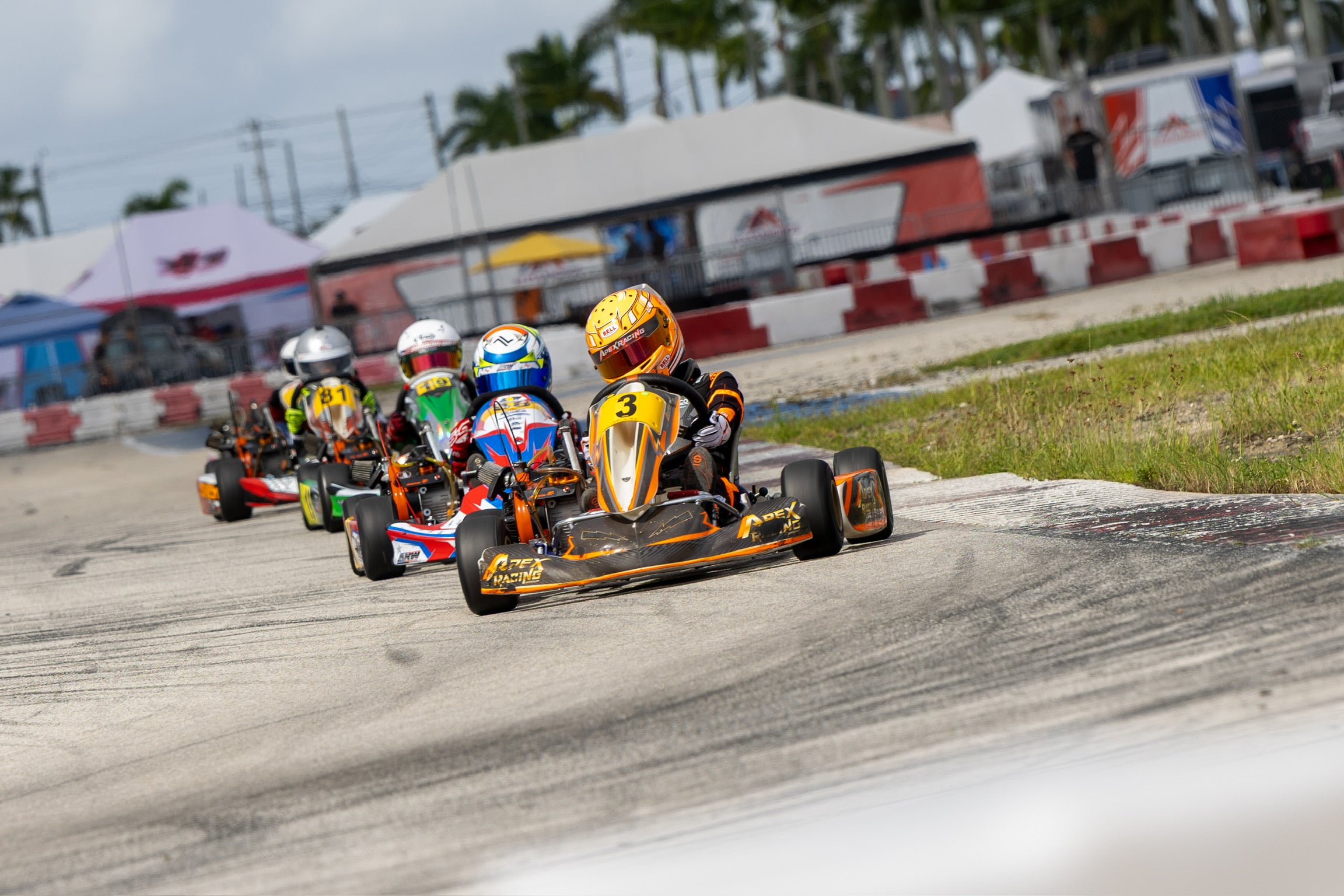 Children racing go-karts on a track with tents and palm trees in the background.